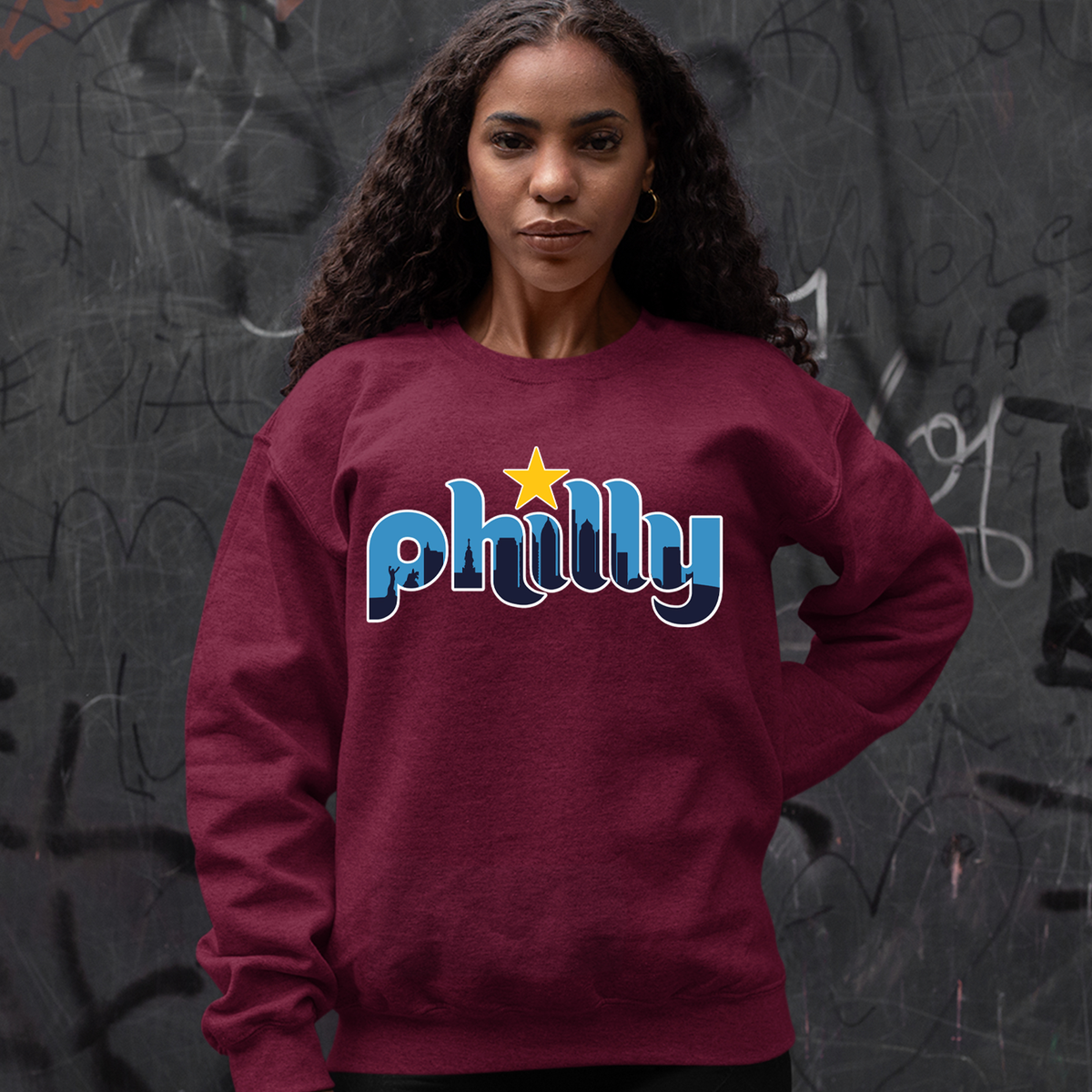 Person wearing a maroon Philly skyline sweatshirt in front of a dark graffiti wall