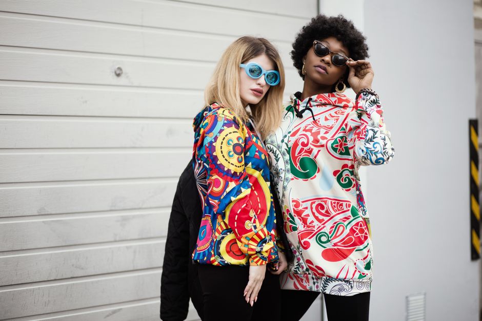 Two stylish women wearing colorful graphic hoodies and sunglasses against a modern urban backdrop.