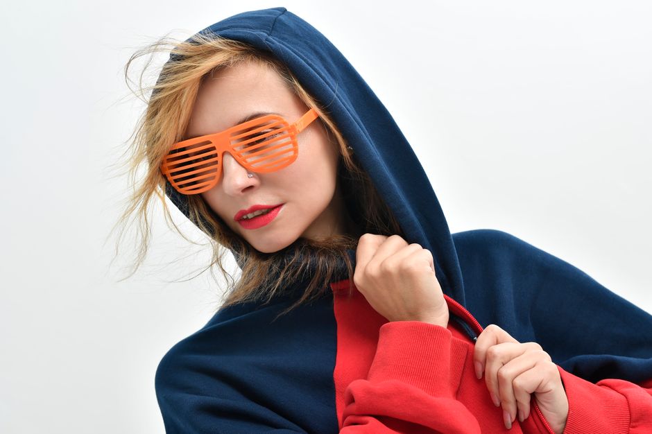 Trendy woman wearing a hoodie and orange sunglasses, striking a pose against a white background.