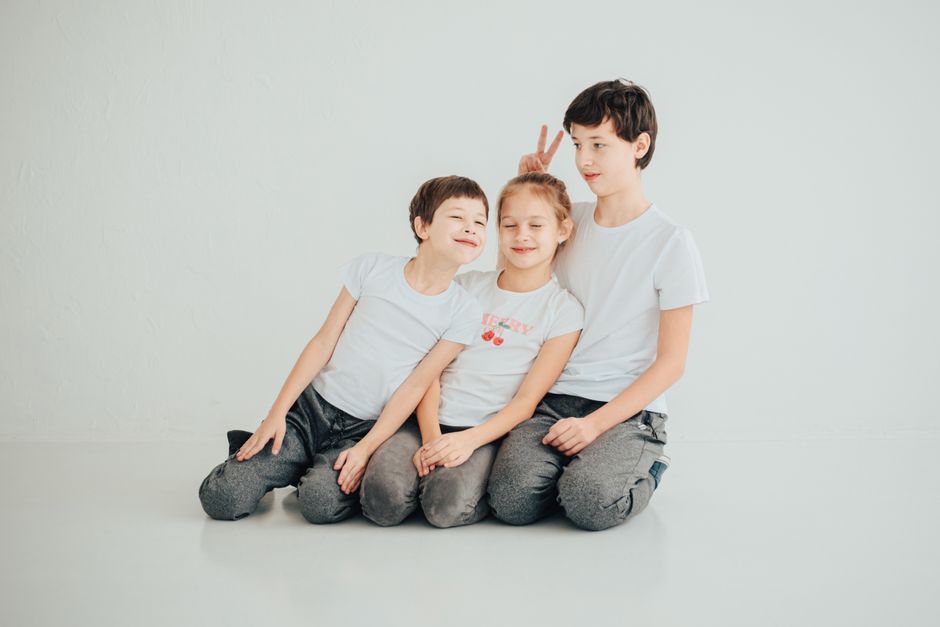 Three siblings playfully pose wearing white t-shirts on a white surface indoors.