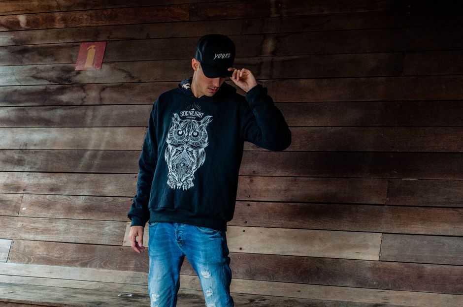 Fashionable man wearing a hoodie with owl design and cap posing against a wooden wall.
