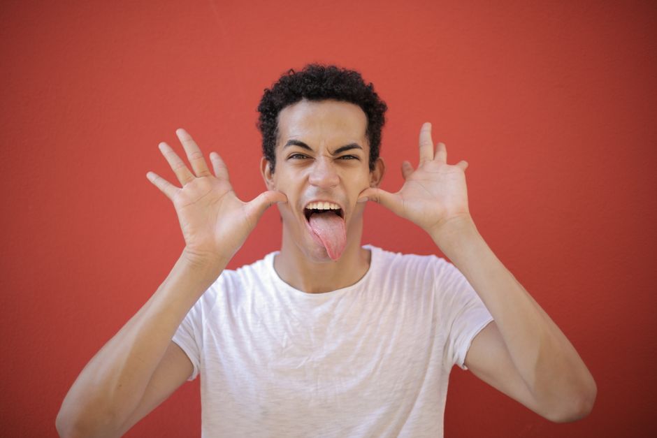 Cheerful young guy with open palms near head sticking out tongue while making grimace against red background